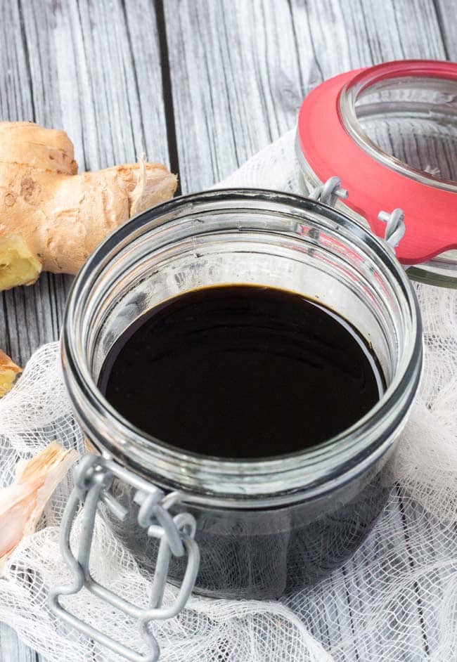 A glass, flip top jar full of healthy teriyaki sauce. The jar sits on a piece of white muslin and a grey background, with a knob of ginger behind the glass jar. 