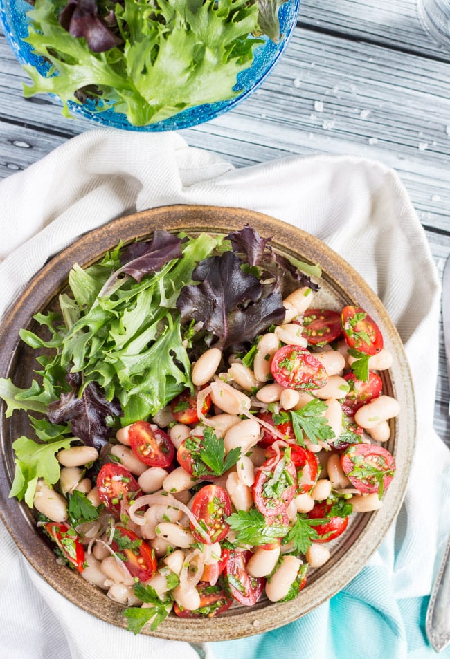 A brown earthenware plate, filled with tomato & white bean salad and green salad leaves.