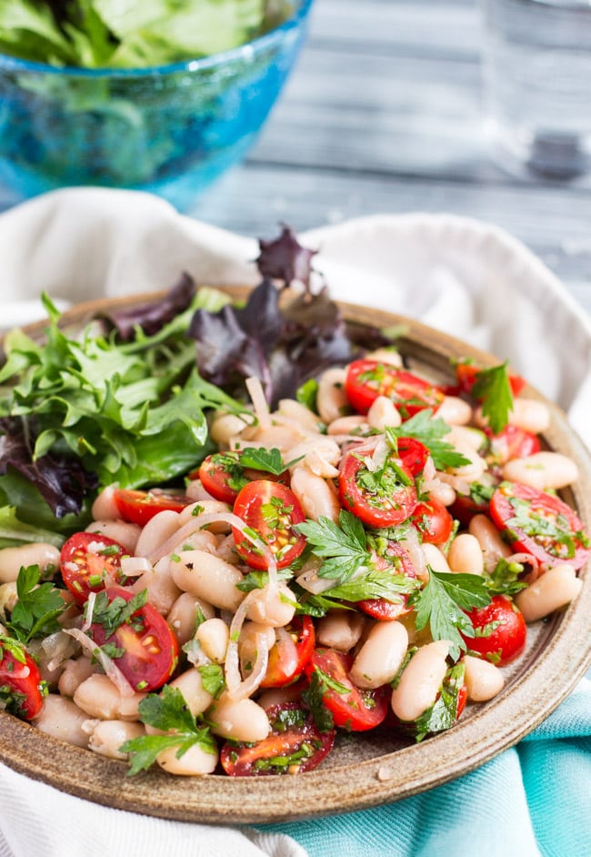 A plateful of tomato & white bean salad, with a blue bowl full of green salad leaves in the background. 